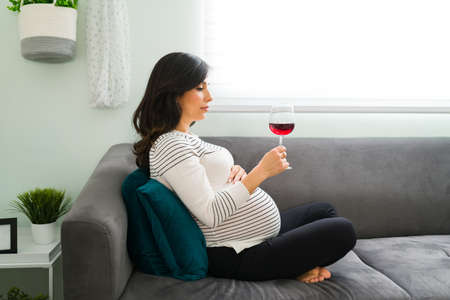 Depressed pregnant woman deciding whether or not to drink a glass of wine while sitting on the sofa. Caucasian expectant mother with bad and unhealthy habitsの写真素材