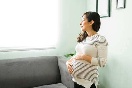 Pregnant woman in her 30s touching and caressing her belly with a reflective and thoughtful expression. Expectant mother looking out the window at homeの写真素材
