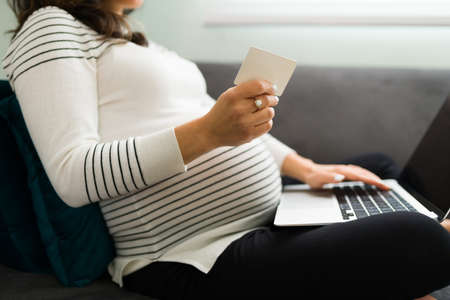 Close up of an expectant mother holding a credit card to buy online baby clothing. Pregnant woman using a laptop at home to purchase stuff for her newborn babyの写真素材