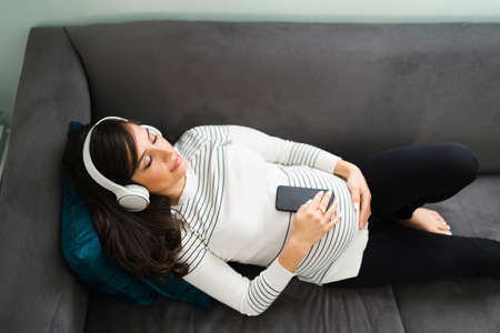Calm pregnant woman seen from above meditating with her eyes closed while lying on the couch. Serene expectant mother listening to meditation musicの写真素材
