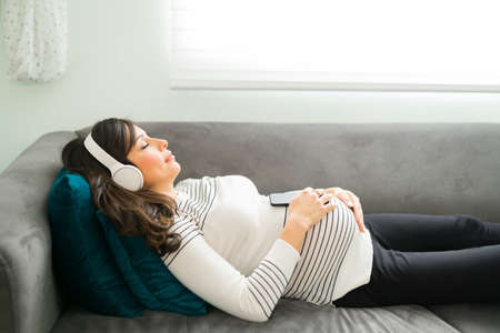 Relaxed pregnant woman lying on the sofa while listening to meditation music. Caucasian pregnant woman meditating with headphones on and her eyes closedの写真素材