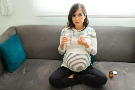 Caucasian pregnant woman looking at the camera with a serious expression while holding a glass of water and a bottle of vitamin pillsの写真素材