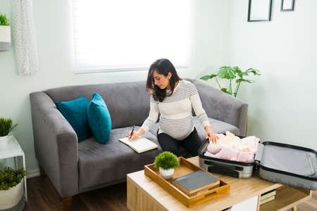 Excited pregnant woman in her 30s planning and writing on her agenda in the living room. Happy expectant mother preparing her hospital suitcase for the birth of her newbornの写真素材