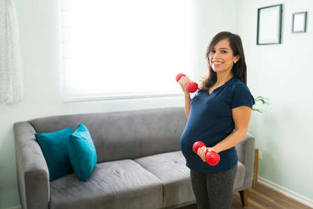 Sporty pregnant woman in sportswear with a big smile lifting weights in her living room. Happy expectant mother exercising at home to stay healthyの写真素材