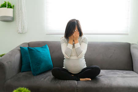 Depressed pregnant woman feeling sad and crying while sitting on the living room sofa. Expectant mother affected by hormones sobbing and covering her faceの写真素材