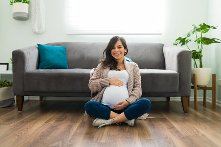 Adorable pregnant woman in her 30s sitting on her living room floor and showing her pretty belly. Attractive expectant mother smiling and posing at homeの写真素材