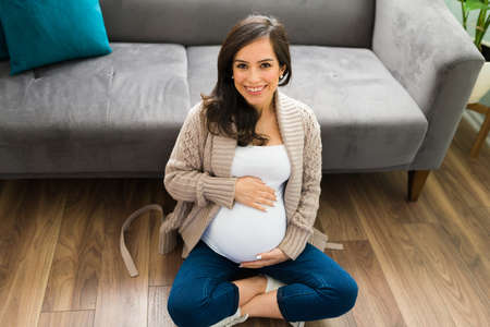 High view of a beautiful pregnant woman sitting with her legs crossed on her living room floor. Proud expectant mother embracing her pretty bellyの写真素材
