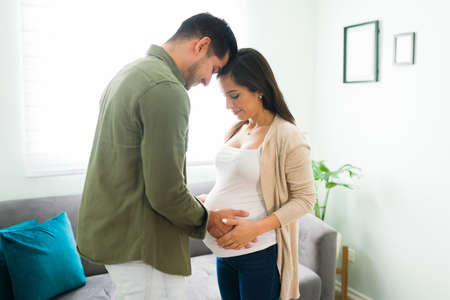 Beautiful couple in love standing in the living room and caressing the belly of an attractive pregnant woman. Latin husband and caucasian woman expecting a baby and holding handsの写真素材