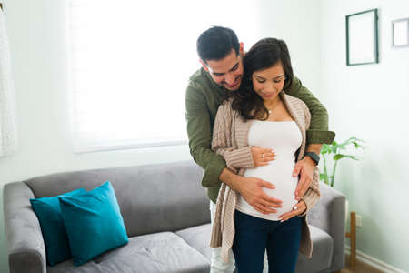 Loving and adorable couple talking to their baby in the belly. Latin man and caucasian pregnant woman in love hugging in the living roomの写真素材