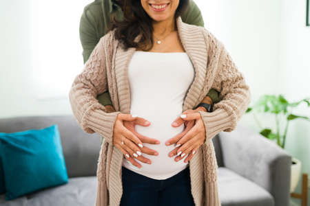 Proud hispanic man hugging a happy pregnant woman from behind. Married couple touching together the belly with their future babyの写真素材