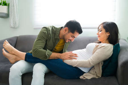 Attractive loving man talking to the belly of a pregnant woman while resting on the couch. Pretty pregnant woman with her feet up in her husband's legsの写真素材