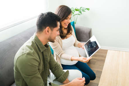Happy pregnant woman and attractive man smiling and holding a tablet with a digital ultrasound on the screen while sitting on her living room sofaの写真素材