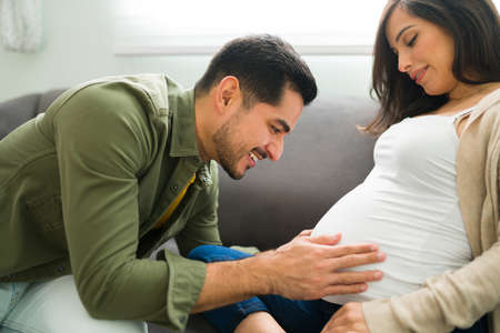 Excited future parents resting on the sofa. Attractive man touching and talking to his pregnant...