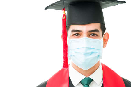 Handsome man in his 20s with a graduation cap and a face mask against a white background. Male young graduate attending his graduation ceremony during the covid pandemicの写真素材