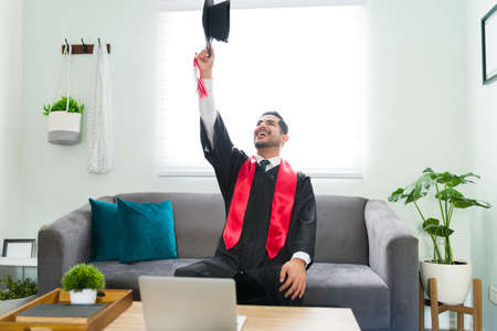 Excited young man throwing a graduation cap in the air while sitting on the living room couch. Thrilled guy in his 20s celebrating during a video call after graduating from universityの販売画像
