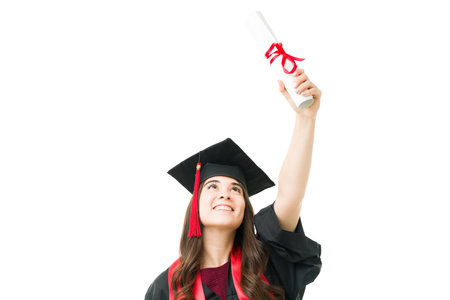Beautiful young college graduate smiling and raising in the air her university diploma while wearing...