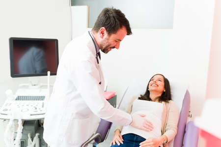 Happy male doctor touching with gloves the belly of a pregnant woman. Female patient smiling while...