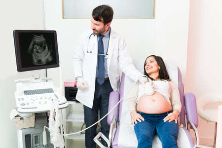 Male gynecologist doing an ultrasound on a pregnant woman. Excited pregnant woman looking at the sonogram of her baby in the machineの写真素材