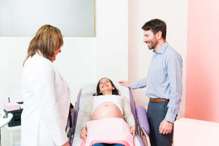 Female gynecologist talking with a patient and her husband about doing an ultrasound. Pregnant woman lying on the examination chair and listening to her doctorの写真素材