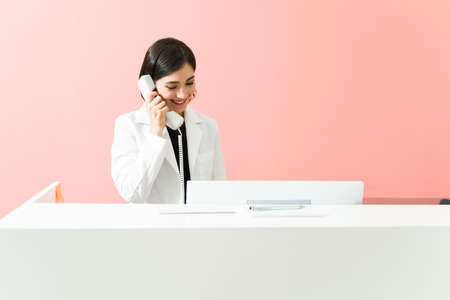 Caucasian receptionist smiling while talking on the phone with a patient. Female worker making a medical appointment at the reception deskの写真素材