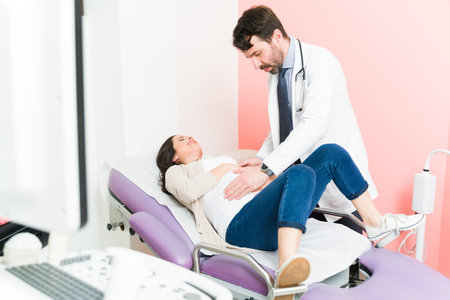 Worried pregnant woman lying at the doctor's chair and complaining about pain in her belly. Male gynecologist examining and touching the stomach of an expectant motherの写真素材