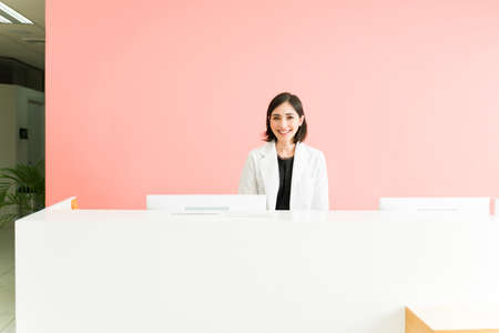 Portrait of a female receptionist in a white robe working at a doctor's office. Caucasian woman smiling and standing behind a deskの写真素材