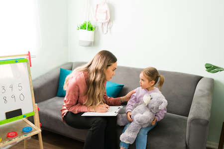 Female therapist touching the arm of a sad elementary girl with a teddy bear. Caucasian psychologist explaining and talking to a little girl about negative feelings and emotionsの写真素材