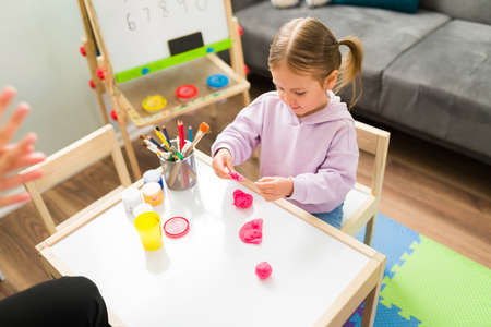 High angle of an adorable girl modeling clay while sitting at a kid's table in her therapist's...