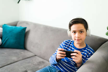 Happy caucasian boy with headphones playing videogames on his smartphone while waiting for his...