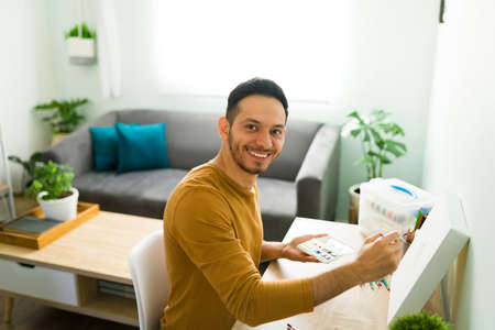 Portrait of a happy man during a leisure day at home. Positive latin man painting a watercolor artwork to keep a good mental healthの写真素材