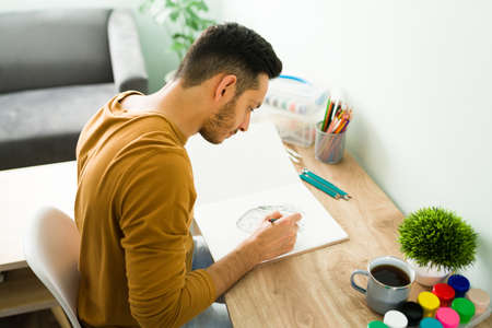 Good-looking man with positive mental health doing a sketching on a white sheet of his notebook while sitting at his deskの写真素材
