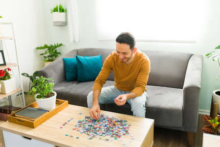 Happy man doing a mental health activity while resting in his living room. Hispanic man putting together a jigsaw puzzle while sitting on the couchの写真素材
