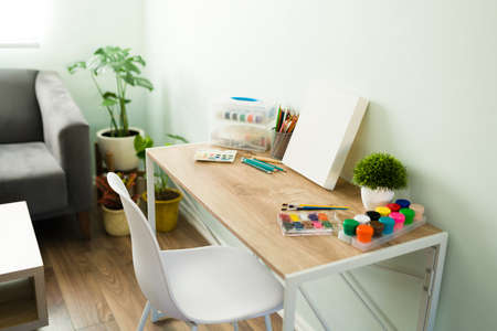 Modern desk and stylish chair in the living room with painting brushes, colored pencils, watercolor paint, ink, a blank canvas and crafting toolsの写真素材