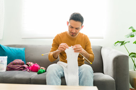 Adult man sitting on the sofa and knitting a scarf as part of his mental health activities. Hispanic man using needles and wool balls to knit clothesの写真素材