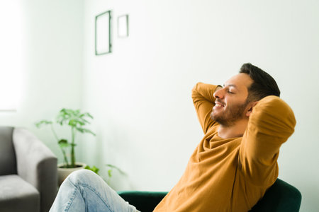 Happy handsome man sitting in his living room. Man with good mental health relaxing with his eyes closed and the hands behind the headの写真素材