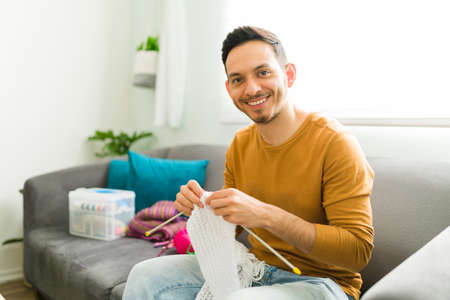 Portrait of a smiling man holding knitting needles and doing a wool scarf as part of his hobby to keep a positive mental healthの写真素材