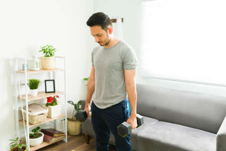 Attractive young man standing in the living room with two weigths and preparing to start his home workoutの写真素材
