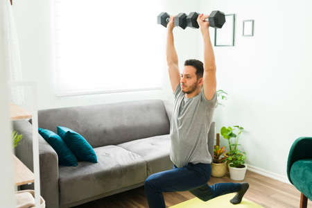 Good-looking man in his 30s doing leg plunges while raising wieghts above his head. Latin man exercising in the living roomの写真素材