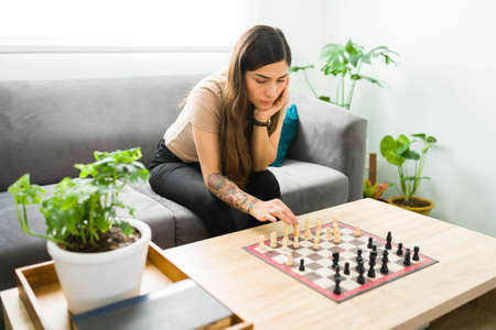 Beautiful hispanic woman moving a chess piece while sitting on the couch. Young woman playing chess alone as a part of her mental health activitiesの写真素材
