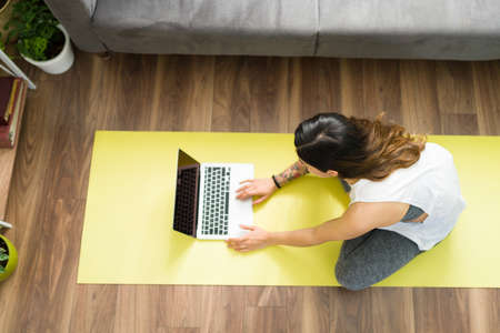 Latin woman searching online in the laptop for a workout video tutorial while sitting on a yoga mat in her living roomの写真素材