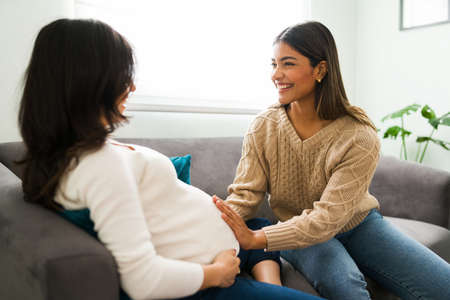 Expectant mother sitting on the sofa during a home visit from her happy doula. Smiling midwife touching the belly of a pregnant womanの写真素材