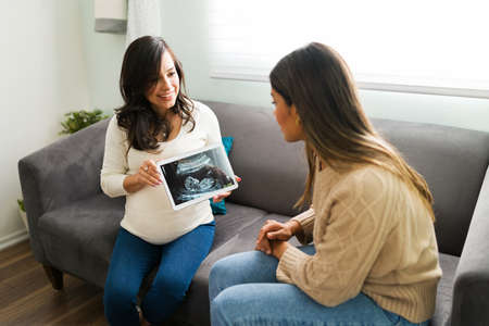 Attractive pregnant woman showing the ultrasound of her baby to her best friend while sitting in the...