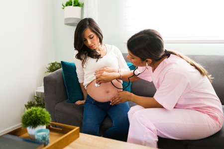 Worried pregnant woman using the medical services of a professional nurse at home. Female doctor listening to the heartbeat of the baby with a stethoscopeの写真素材