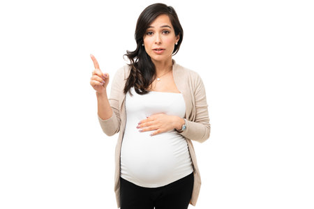 Caucasian pregnant woman pointing up with her finger to copy space while talking about  pregnancy tips. Attractive pregnant woman giving advice while touching her bellyの写真素材