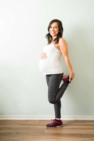 Excited pregnant woman wearing sportswear and standing in one leg. Pretty pregnant woman stretching her legs to start her home workoutの写真素材