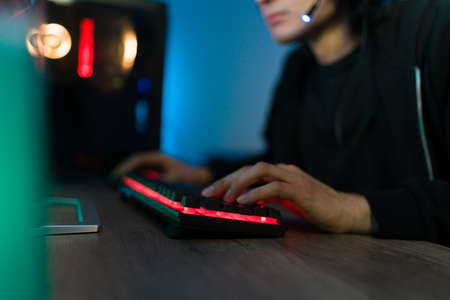 Hispanic young man pushing buttons and using a gaming keyboard with led lights to play an online...