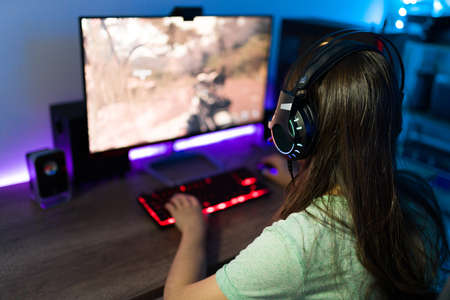 Woman gamer with headphones seen from behind while playing a video game in her gaming PC with neon...