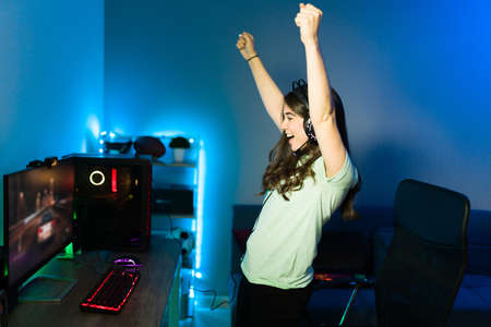 Attractive young woman cheering and screaming while standing in front of her desk with a gaming computer PC. Female gamer winning an online video gameの写真素材