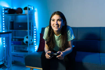 Young female gamer having fun and using a remote controller to play a video game while sitting on the sofa in her bedroom decorated with led lightsの写真素材