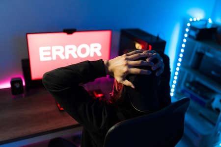 Young man seen from behind holding his head and looking surprised while watching an error message on a gaming PC after playing an online videogameの写真素材